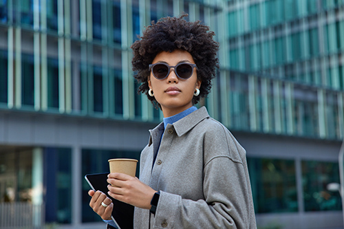 Fashionable young businesswoman dressed in grey jacket drinks coffee from paper disposable cup holds modern tablet poses against modern business building outside wears sunglasses looks away.